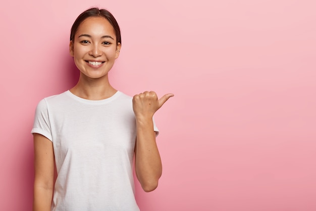 Horizontal shot of happy young Asian woman points away on copy space, demonstrates something good, wears white t shirt, helps pick best choice, recommends product, models over pink wall