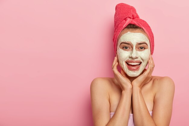 Горизонтальный снимок счастливой молодой европейской девушки, которая накладывает грязевую маску на лицо, трогает щеки, избегает раздражения кожи, имеет обнаженные плечи.