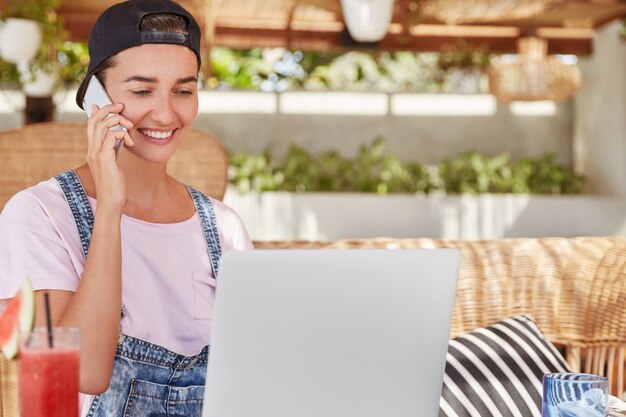 Горизонтальный снимок счастливой молодой женщины в стильной кепке радостно смотрит на экран ноутбука, делает онлайн-проект, подключенный к беспроводному Интернету, разговаривает с кем-то на смартфоне.