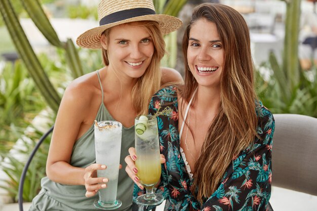 Горизонтальный снимок счастливых молодых женщин, которые наслаждаются летним отдыхом, чокаются с коктейлями, сидят вместе в кафе на террасе. Гомосексуальная пара приходит на вечеринку, рада общению, свободное время