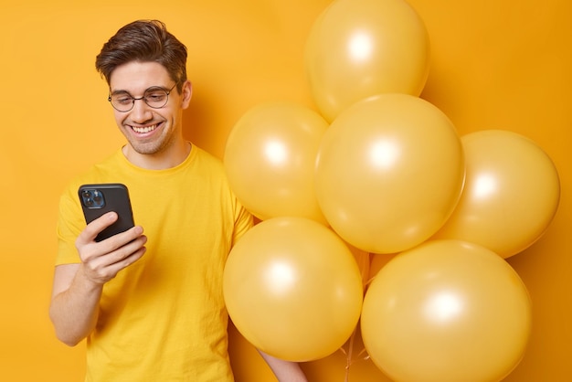 행복한 청년이 스마트폰에서 생일을 축하하는 알림을 확인하는 수평 사진은 노란색 배경 위에 풍선이 격리된 캐주얼한 티셔츠를 입고 파티에 나옵니다.