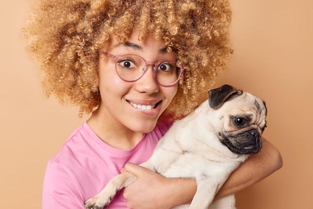 Horizontal shot of happy young woman carries pug dog loves animals carries pet to veterinary clinic wears spectacles casual t shirt isolated over brown background Look at my new family member