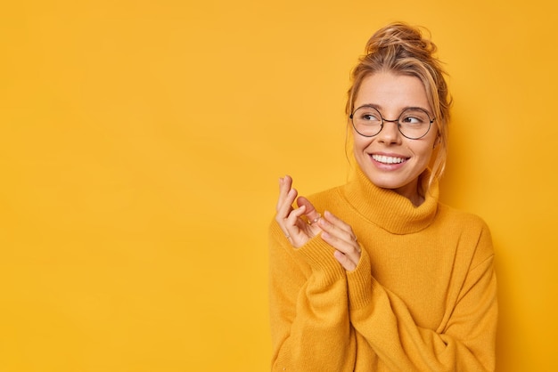 Free photo horizontal shot of happy young woman rubs hands looks curious awayy smiles toothily wears casual jumper and round spectacles isolated over yellow background blank space for your advertising content