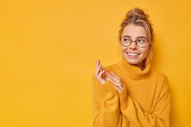 Горизонтальный снимок счастливой молодой женщины, потирающей руки, выглядит любопытно, зубасто улыбается, носит случайный джемпер и круглые очки, изолированные на желтом фоне, пустое место для вашего рекламного контента