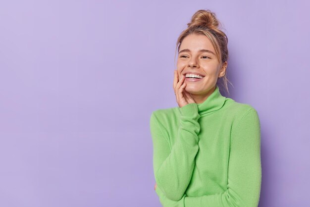 Горизонтальный снимок счастливой молодой женщины, улыбающейся зубасто, смотрит куда-то, чувствует себя радостным, думает о чем-то приятном, носит повседневную зеленую водолазку, изолированную на фиолетовом фоне с копией пространства.