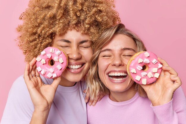 Horizontal shot of happy young women hold delicious doughnuts with marshmallow have tasty snack smile broadly keep eyes closed have good mood while eating dessert pose indoor Sugar addiction