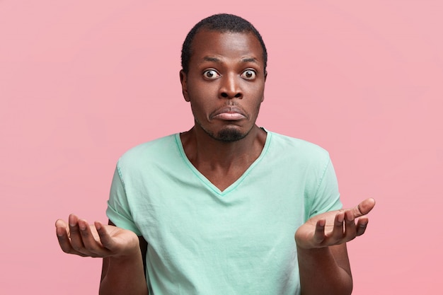 Free photo horizontal shot of hesitant handsome dark skinned male shrugs shoulders, being uncertain and unsure, curves lower lip and stares, poses against pink