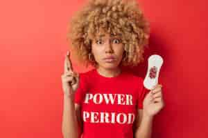 Free photo horizontal shot of hopeful european woman holds dirty sanitary napkin keeps fingers crossed hopes to get pregnant next time has worried expression isolated over red background blood period