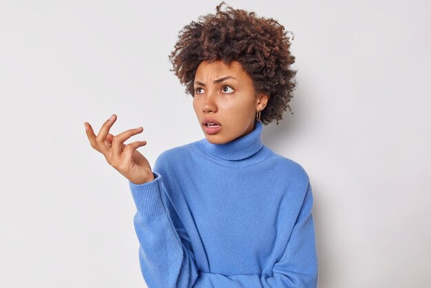 Horizontal shot of indignant woman raises palm feels questioned displeased wears blue turtleneck puzzled by unexpected news isolated over white background gestures skeptically doesnt know what to do