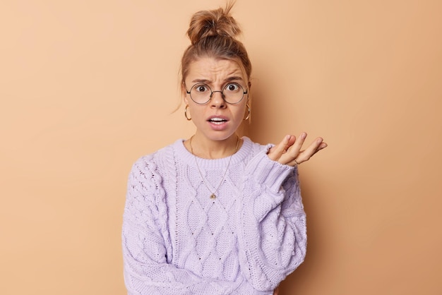 Horizontal shot of indignant young European woman with combed hair looks puzzled shrugs shoulders feels confused wears round spectacles and casual jumper isolated over beige background So what