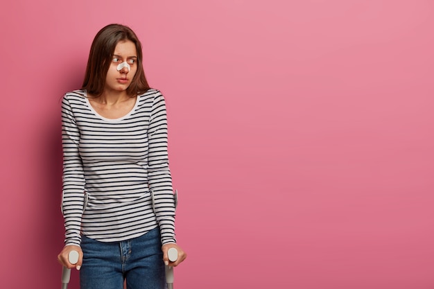 Horizontal shot of injured woman got into car accident, recovers at home, has serious trauma, looks aside, wears medical plaster on broken nose, isolated on pink wall. Disability, medical care concept