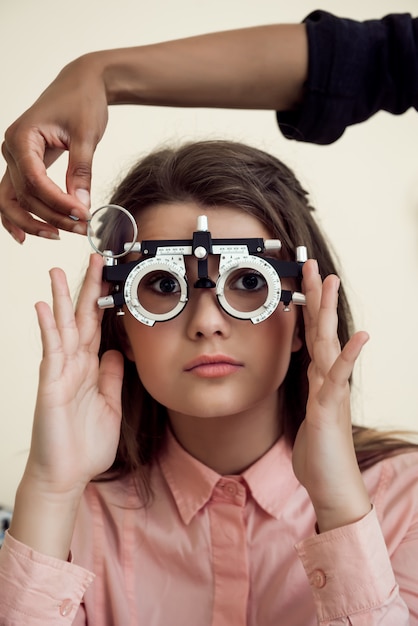 Horizontal shot of interested and curious caucasian girl on appointment with eye care specialist wearing phoropter while ophthalmologist checking her vision, sitting over yellow wall