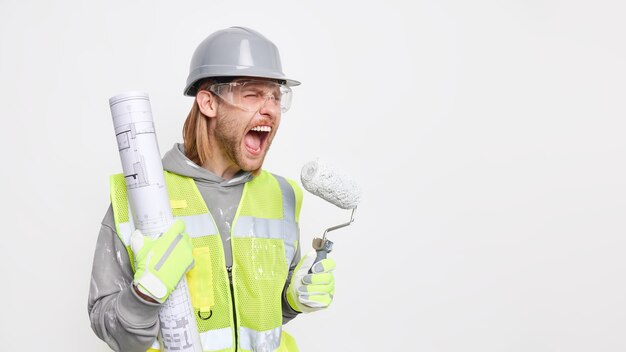 Horizontal shot of irritated male construction worker exclaims negatively holds blueprint and painting roller