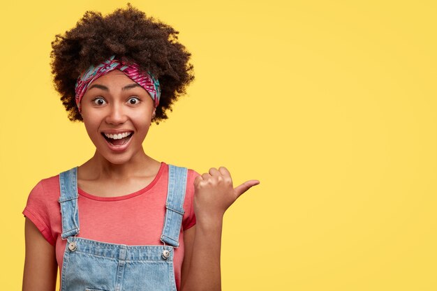 Horizontal shot of joyful curly dark skinned African American female with overjoyed expression, points aside against yellow wall, dressed in fashionable dungarees