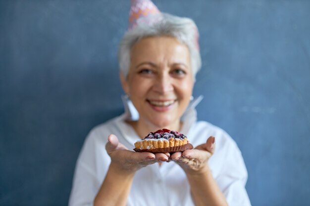 Horizontal shot of joyful positive mature female with gray hair enjoying birthday party, eating blackberry pie. Selective focus on cake