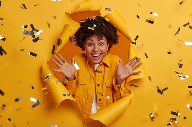 Free photo horizontal shot of joyous woman with afro hairstyle, glad to see close person, raises palms, wears stylish outfit excited to pose in paper hole with flying sparkles