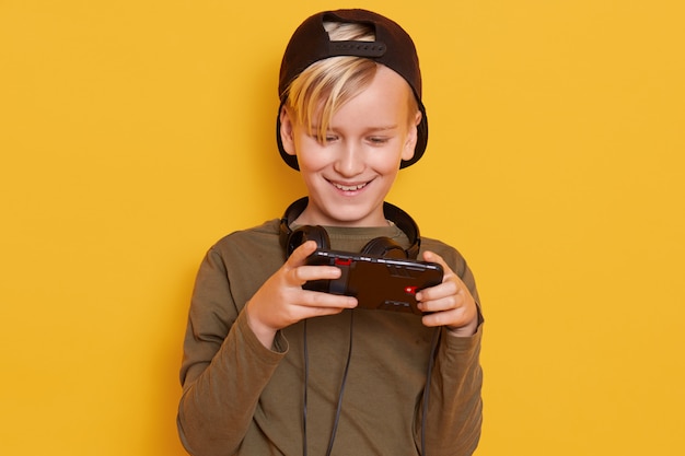 Horizontal shot of little boy wearing black cap and green hoody, posing with cell phone in hands, fashionable kid playing online games.