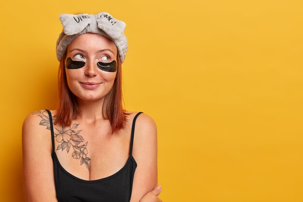 Horizontal shot of lovely adult woman dressed in sleevless black t shirt, has tattoo on body, looks away with thoughtful expression, wears headband and hydrogel patches, cares about skin and beauty