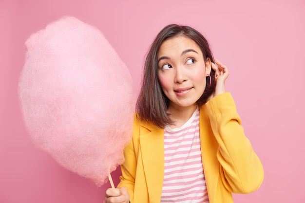 Horizontal shot of lovely brunette Asian teenage girl looks away with thoughtful expression holds delicious cotton candy wears yellow jacket isolated over pink wall. Sweet tooth concept