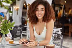 Free photo horizontal shot of lovely dark skinned female model with curly afro hairdo, enjoys recreation time during weekend, poses agianst cozy cafe interior