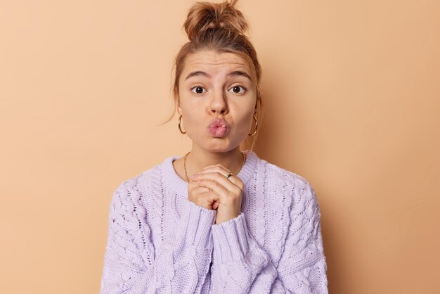 Horizontal shot of lovely young woman keeps lips folded feels romantic keeps hands together wants to kiss someone looks with tender expression dressed in knitted jumper isolated over beige wall