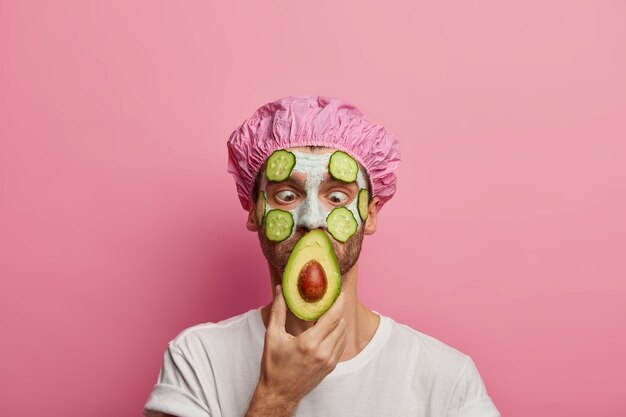 Horizontal shot of male looks with bugged eyes at acovado, wears bath cap, applies beauty facial mask