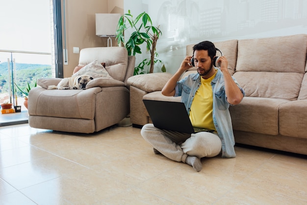 Горизонтальный снимок мужчины, сидящего на полу, слушающего музыку и работающего с ноутбуком дома