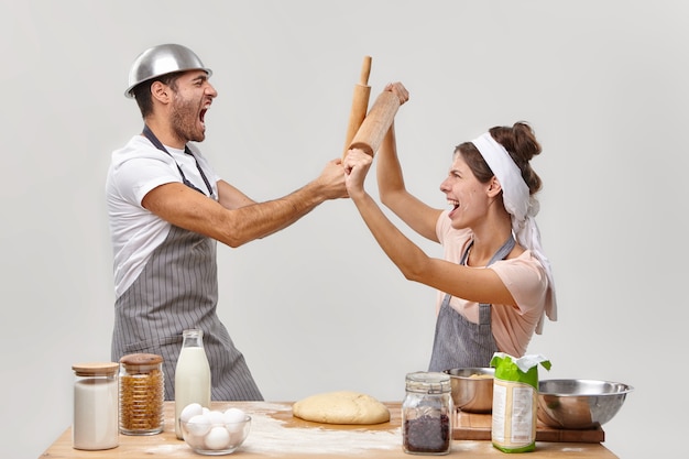 Horizontal shot of man and woman opponents participate in cooking challenge, fight with wooden rolling pins, have culinary battle, work in bakery, make dough, pose at kitchen against white wall