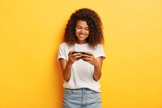 Horizontal shot of millennial young woman with curly hair posing with her phone