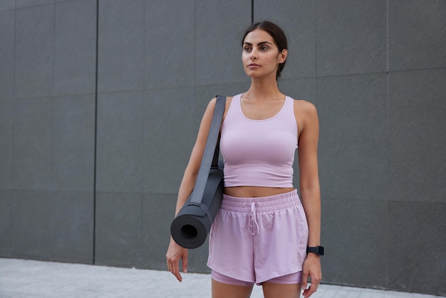 Free photo horizontal shot of motivated slim woman dressed in sportswear prepares for hard training poses with fitness mat focused into distance poses against grey wall with mock up space for your text