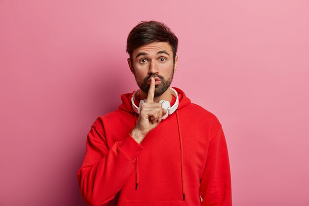 Horizontal shot of mysterious bearded man makes shush gesture, shows hush sign, asks not tell secret, presses index finger to lips, wears red sweater, poses over rosy pastel wall. Secrecy concept