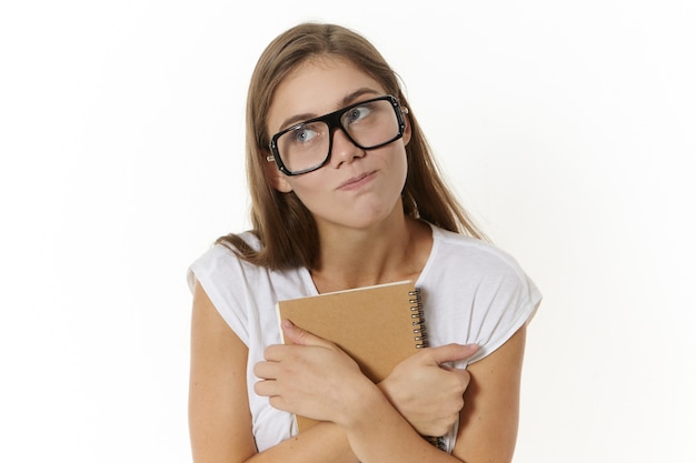 Horizontal shot of mysterious beautiful young woman in eyewear looking away and embracing copybook or diary, trying to hide something interesting that her wrote down, does not want to share her secret