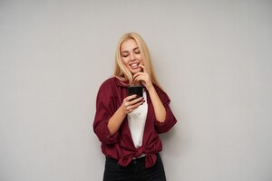 Бесплатное фото Горизонтальный снимок привлекательной позитивной блондинки с распущенными волосами, которая весело смотрит на экран своего смартфона и искренне улыбается, прижимая указательный палец к губе