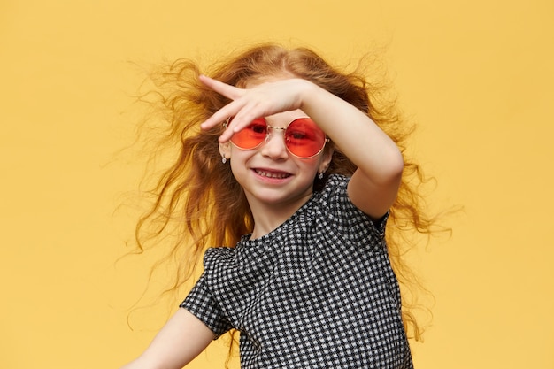 Горизонтальный снимок красивой счастливой модной маленькой девочки с вьющимися рыжими волосами, наслаждающейся танцами, с веселой широкой улыбкой, в солнечных очках. музыка, танцы, развлечения и детская концепция