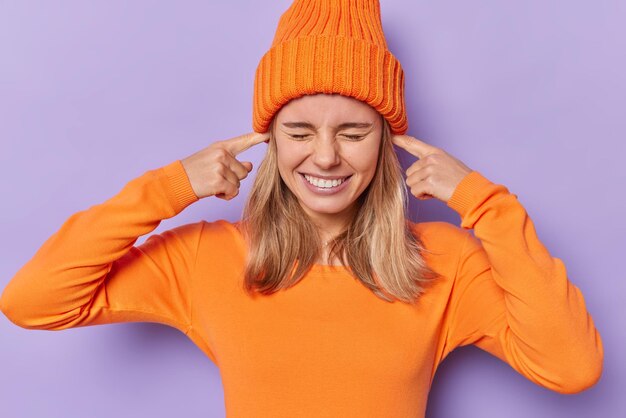 Горизонтальный снимок эмоциональной светловолосой молодой женщины, затыкающей уши пальцами, избегающей громкой музыки или шума, с закрытыми глазами, в повседневном оранжевом джемпере с длинными рукавами и шляпе, изолированной на фиолетовом фоне