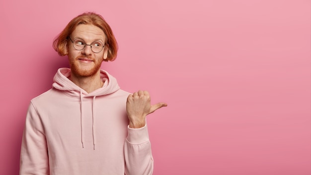 Бесплатное фото Горизонтальный снимок рыжего бородатого кавказца показывает большим пальцем вправо, в прозрачных очках и толстовке с капюшоном, с интересом демонстрирует пустое пространство, демонстрирует красивую рекламу, изолированную на розовой стене