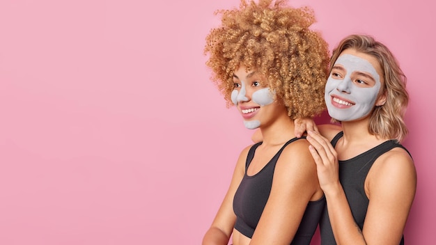 Бесплатное фото Горизонтальный снимок счастливых молодых женщин, стоящих близко друг к другу, питательная маска красоты на лице, улыбка, нежно сфокусированная вперед, изолированная на розовом фоне, пустое место для текста или рекламы