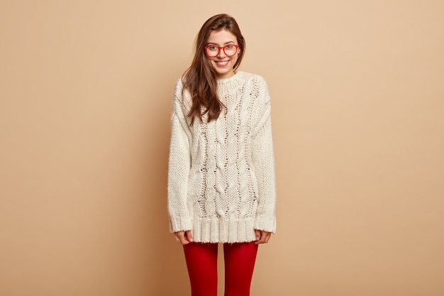 Бесплатное фото Горизонтальный снимок позитивной европейской женщины в белой зимней одежде и красных колготках, которая позирует над бежевой стеной, наслаждается свободным временем и находится в хорошем настроении. люди, эмоции, выражения лица