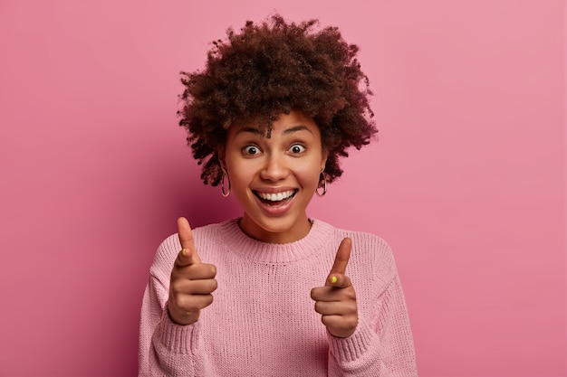 Бесплатное фото Горизонтальный снимок довольно радостной женщины прямо указывает, предлагает присоединиться к ней, выбирает вас, широко улыбается, одетая в повседневный джемпер, позирует на розовой стене. люди и концепция выбора