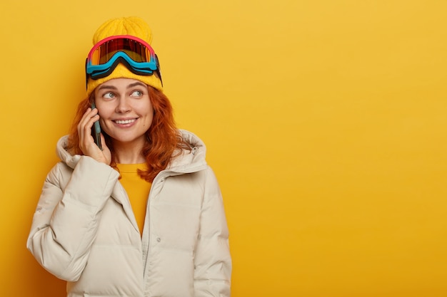 꽤 웃는 여자 스키어의 가로 샷은 친척 마주 스마트 폰을 호출하고, 그녀의 겨울 방학에 대해 이야기하고, 스노우 보드 고글을 착용합니다.
