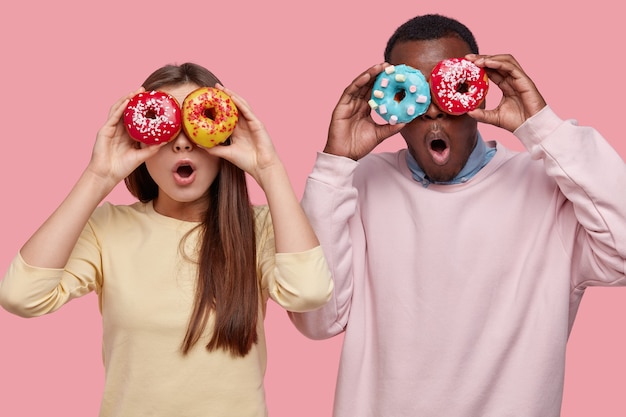 Горизонтальный снимок удивленного смуглого мужчины, прикрывающего глаза вкусными сверкающими пончиками, стоящего рядом с девушкой, вместе проводящего свободное время Бесплатные Фотографии