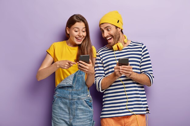 Горизонтальный снимок оптимистичной радостной пары, болтающей с подписчиками из веб-блога, радостно глядя на смартфон