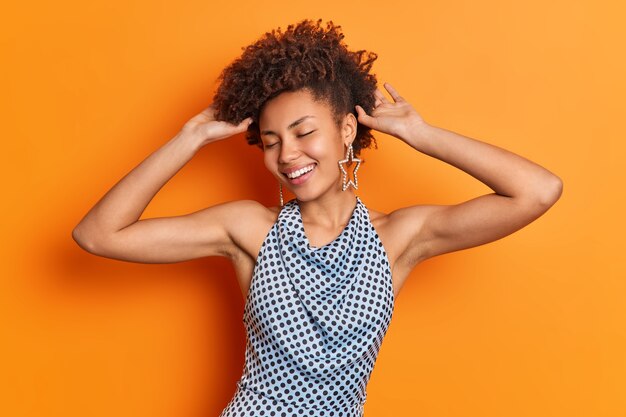 낙관적 인 아프리카 계 미국인 여자의 수평 샷은 곱슬 머리에 손을 유지하고 광범위하게 만족스러운 미소로 눈을 감습니다.