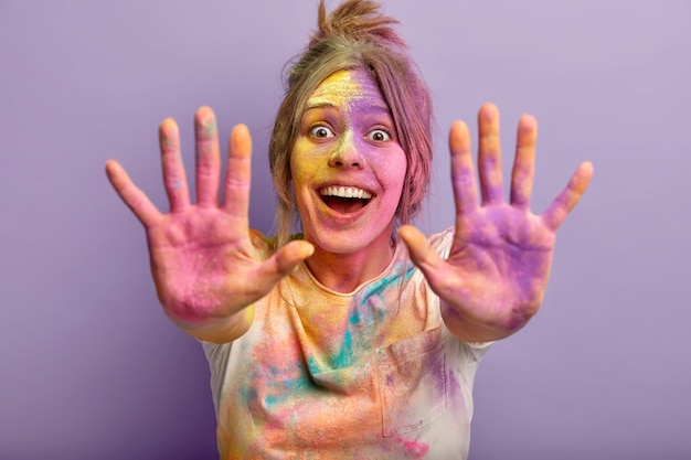 Horizontal shot of optimistic joyful young girl shows two colorful palms, celebrates holi festival, laughs gladfully, plays with special colored powder. Focus on painted hands. Splash of color