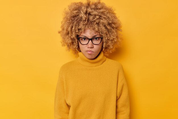 Horizontal shot of outraged curly haired woman frowns face looks angrily being irritated with someone wears transparent glasses and casual sweater isolated over yellow background. Negative emotions