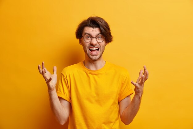 Horizontal shot of peevish angry European man gestures actively exclaims from anger yells with frustrated expression wears casual yellow t shirt expresses negative emotions. Outraged guy screams