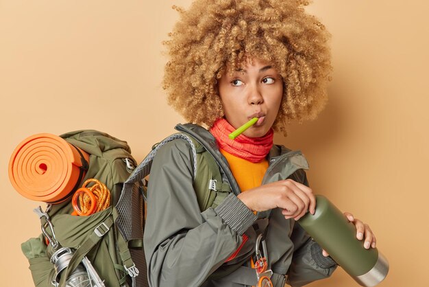 Horizontal shot of pensive curly haired young woman keeps toothbrush in mouth holds thermos with drink dressed in jacket enjoys hiking time poses against brown background Camping trip concept