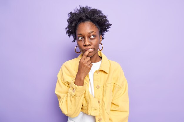 Горизонтальный снимок задумчивой темнокожей молодой женщины, которая делает выбор, держит указательный палец на губах и глубоко о чем-то думает