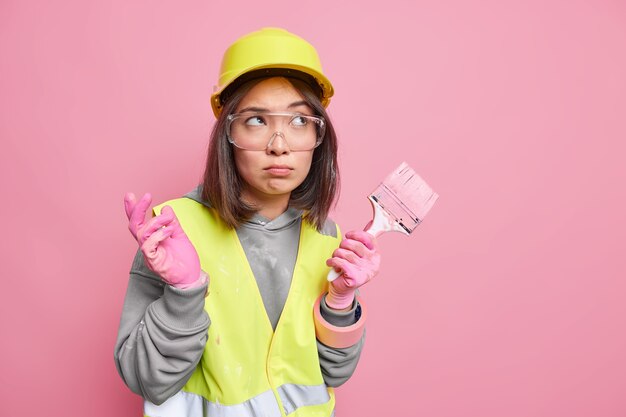 制服を着た物思いにふける女性ビルダーの水平方向のショットは、思考の深いペイントブラシを保持します保護ヘルメット透明メガネ反射ジャケットを着用します