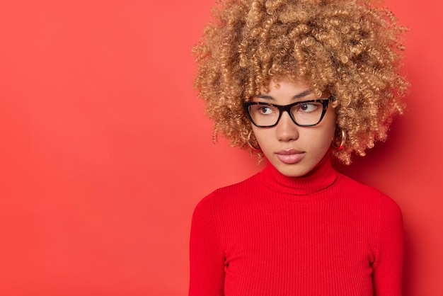 Free photo horizontal shot of pensive woman with curly hair focused away being deep in thiughts wears optical glasses casual poloneck isolated over red background copy space for your promotional content